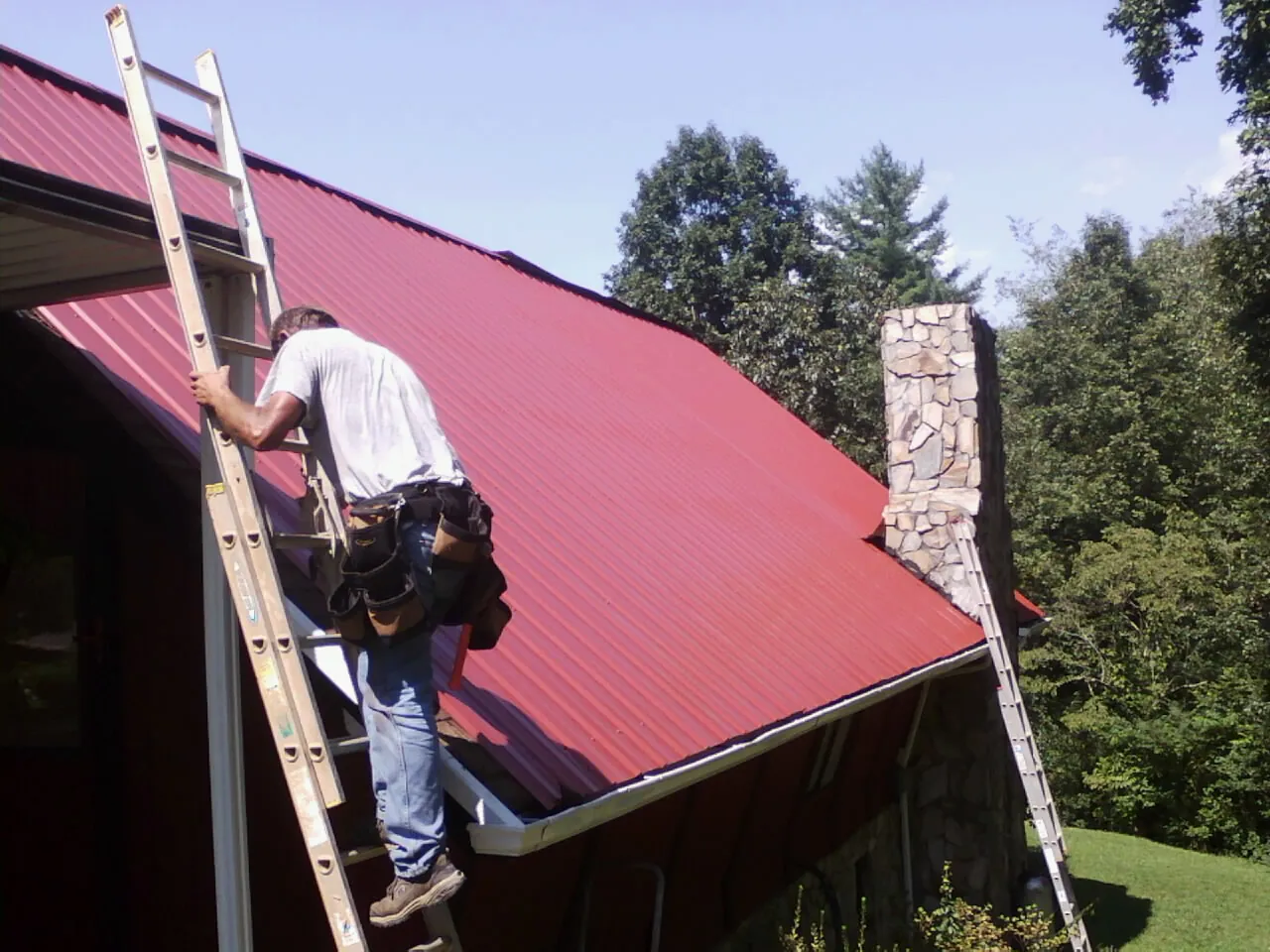 Roofer on a ladder beside a red metal roof and stone chimney — Soffit & Fascia Repair by Roofing Brothers in Wapanucka